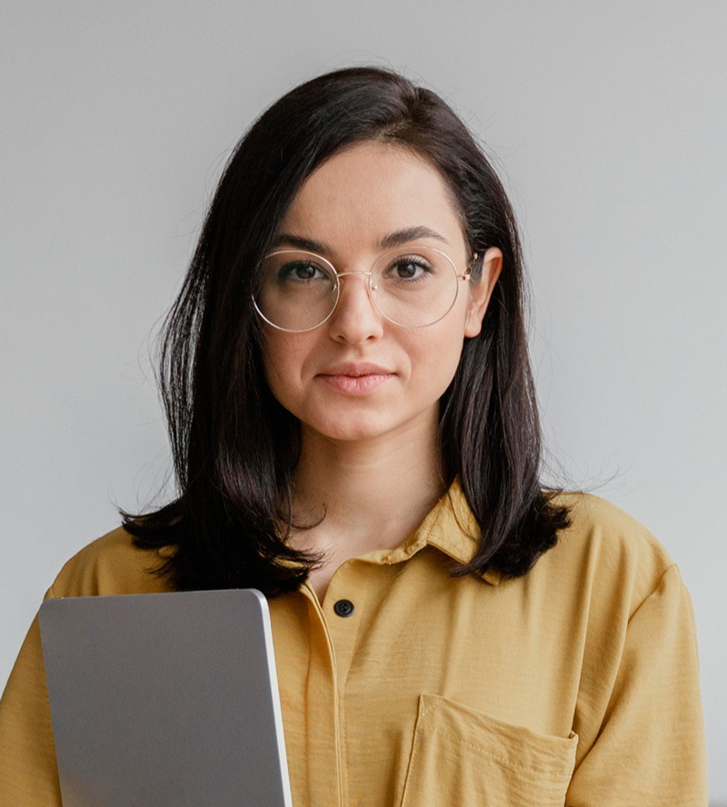 A woman with shoulder-length dark hair and glasses, wearing a yellow shirt, holds a laptop and looks at the camera with a subtle smile.