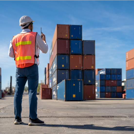 A worker in a safety vest and helmet uses a walkie-talkie in a shipping container yard under a clear blue sky.