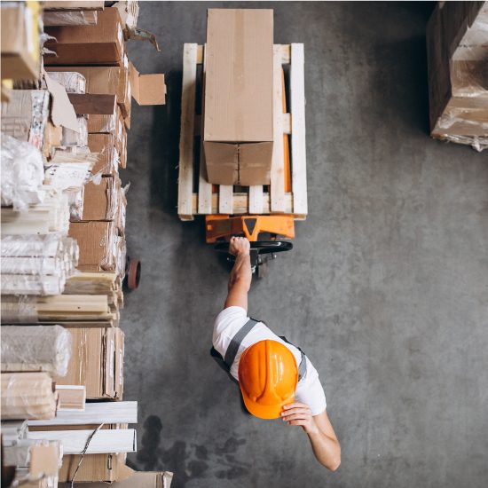 A worker in a hard hat pushing a pallet jack loaded with boxes in a warehouse, viewed from above.