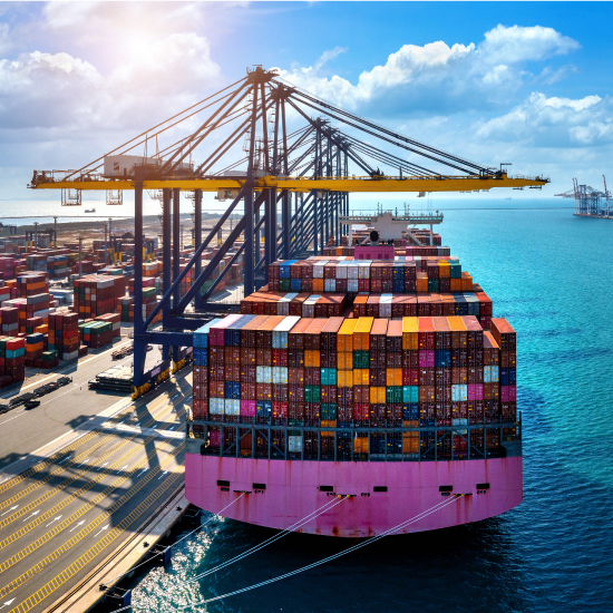 Aerial view of a pink cargo ship loaded with colorful containers at a bustling port, with cranes and clear skies.