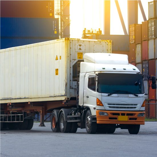 White cargo truck transporting a container in an industrial shipping area during sunset.