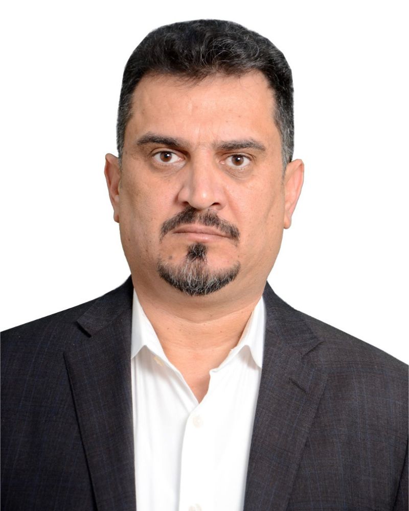 Odai Rashak - Middle-aged man with dark hair and a mustache, wearing a dark suit and white shirt, looking directly at the camera with a serious expression, against a white background.